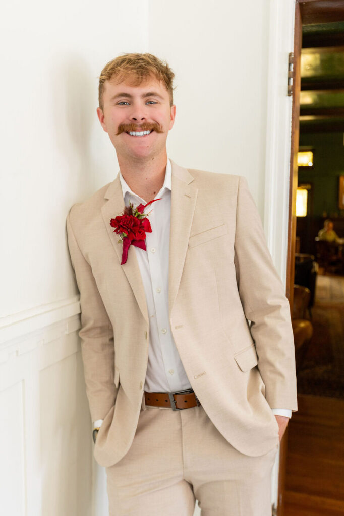 Groom is leaning against a white wall. He is smiling into the camera.